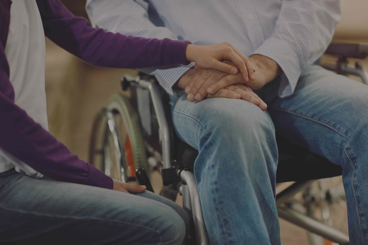 A man and a woman sit next to each other. The man is in a wheelchair. The woman is holding the man's hand.